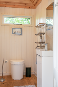 A toilet installed in a Garden Room by Crane Compact bathroom with tongue-and-groove walls, wooden floor, high-level window, toilet and modern white basin.
